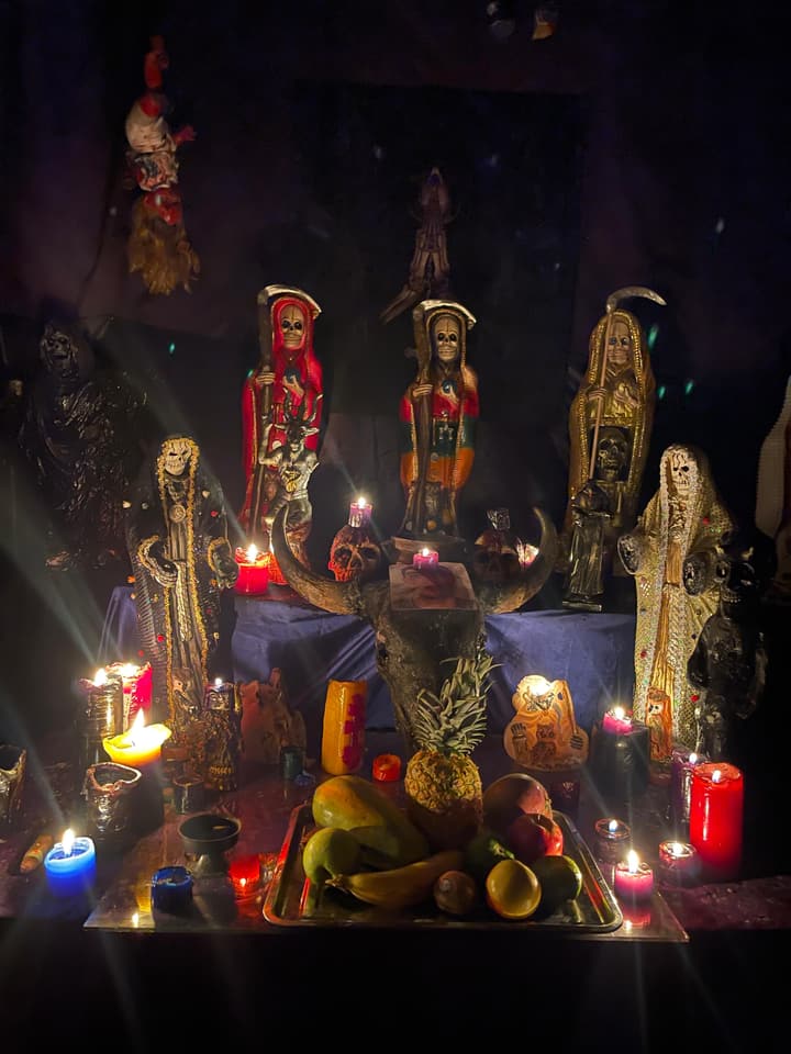 Mi altar de la Santa Muerte: figuras de la Niña Blanca de varios colores, ofrenda de frutas y veladoras encendidas.